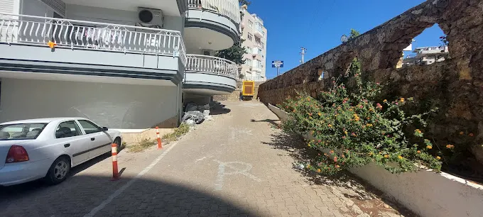 منظر خارجي للمبنى مع موقف سيارات وممر. مشهد خارجي يظهر سيارة بيضاء متوقفة في منطقة مرصوفة بجوار مبنى متعدد الطوابق مع شرفات. جدار حجري مع شجيرات مزهرة يحد ممراً تحت سماء زرقاء صافية.