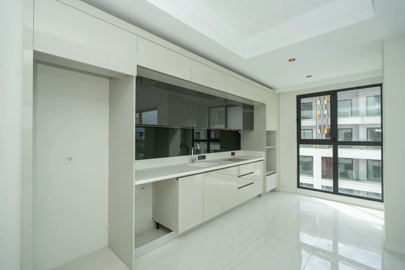 مطبخ أبيض حديث بتصميم أنيق ونافذة كبيرة. مطبخ حديث تم تجديده حديثًا بخزائن بيضاء لامعة وسطح عمل أنيق وأرضيات بلاط لامعة. توفر نافذة كبيرة بإطار أسود إضاءة طبيعية وافرة لهذا الفضاء المشرق.