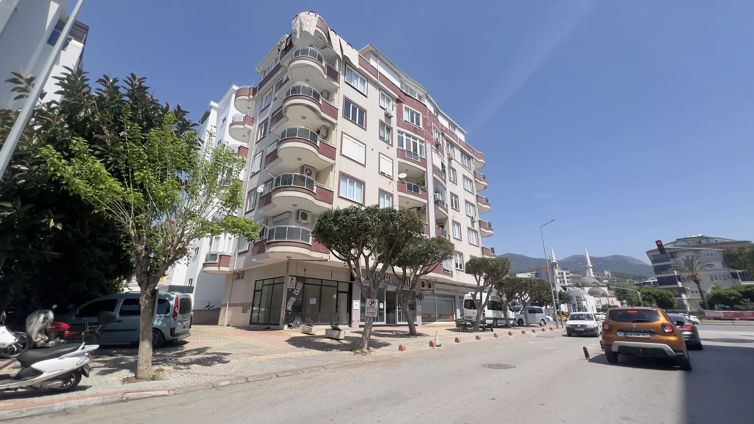 مبنى سكني حديث بشرفات في وسط المدينة. واجهة مبنى سكني حديث متعدد الطوابق مع العديد من الشرفات وطابق أرضي تجاري، يقع في شارع مدينة مشمس به أشجار وسيارات ومسجد في المسافة.