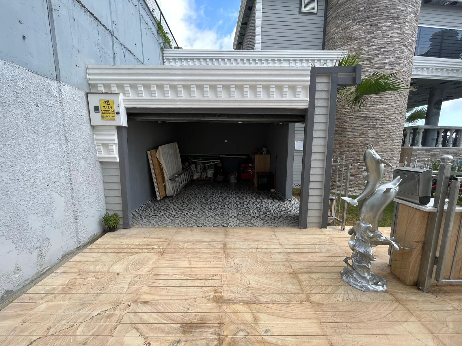مرآب سيارات آمن وتصميم خارجي أنيق للعقار. منظر خارجي لعقار حديث يتميز بمرآب سيارات مفتوح وآمن مع بلاط مزخرف وممر سيارات من البلاط فاتح اللون. يضيف تمثال دلفين مزخرف لمسة من الأناقة.