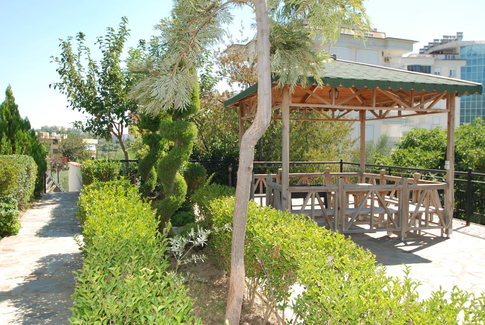 Beautiful Garden Gazebo with Outdoor Seating Area. A well-maintained garden featuring a traditional wooden gazebo with a green roof and integrated seating, surrounded by lush green bushes and trees, offering an ideal outdoor social area.