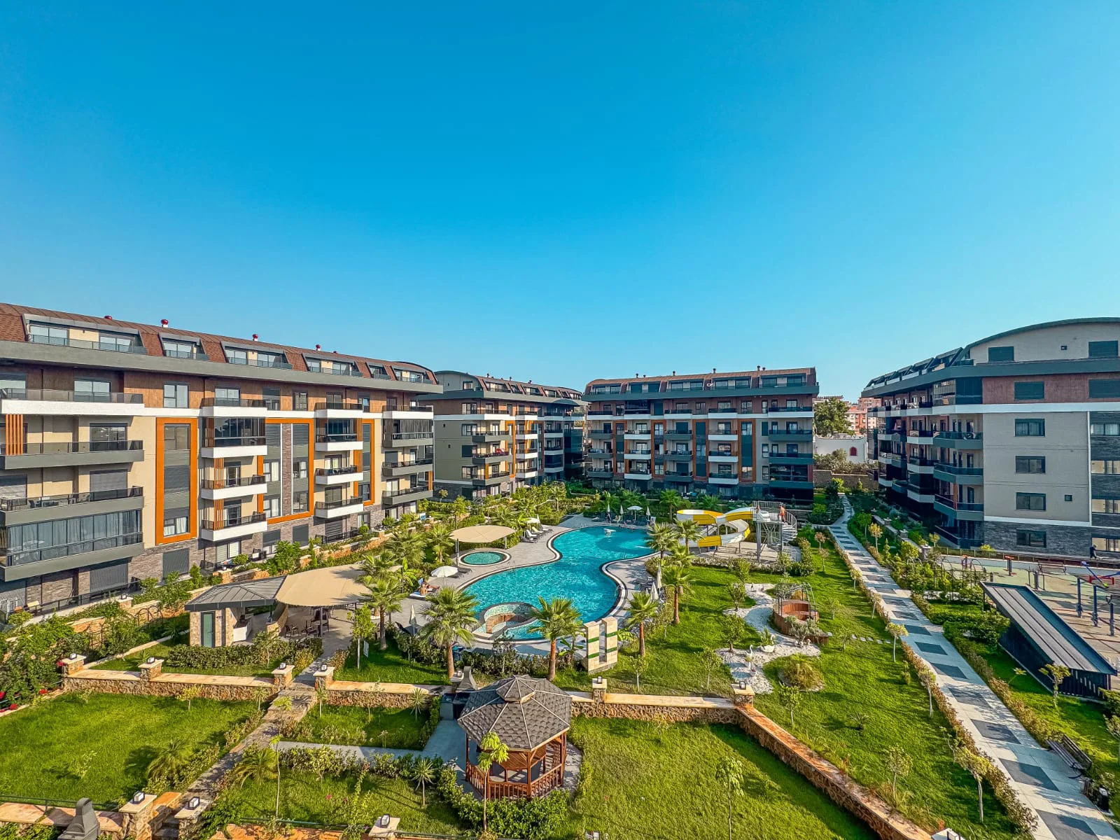 Modern Apartment Complex with Large Outdoor Pool and Gardens. Aerial view of a modern apartment complex featuring multiple buildings, a large resort-style swimming pool, lush landscaped gardens, and a playground under a clear blue sky.