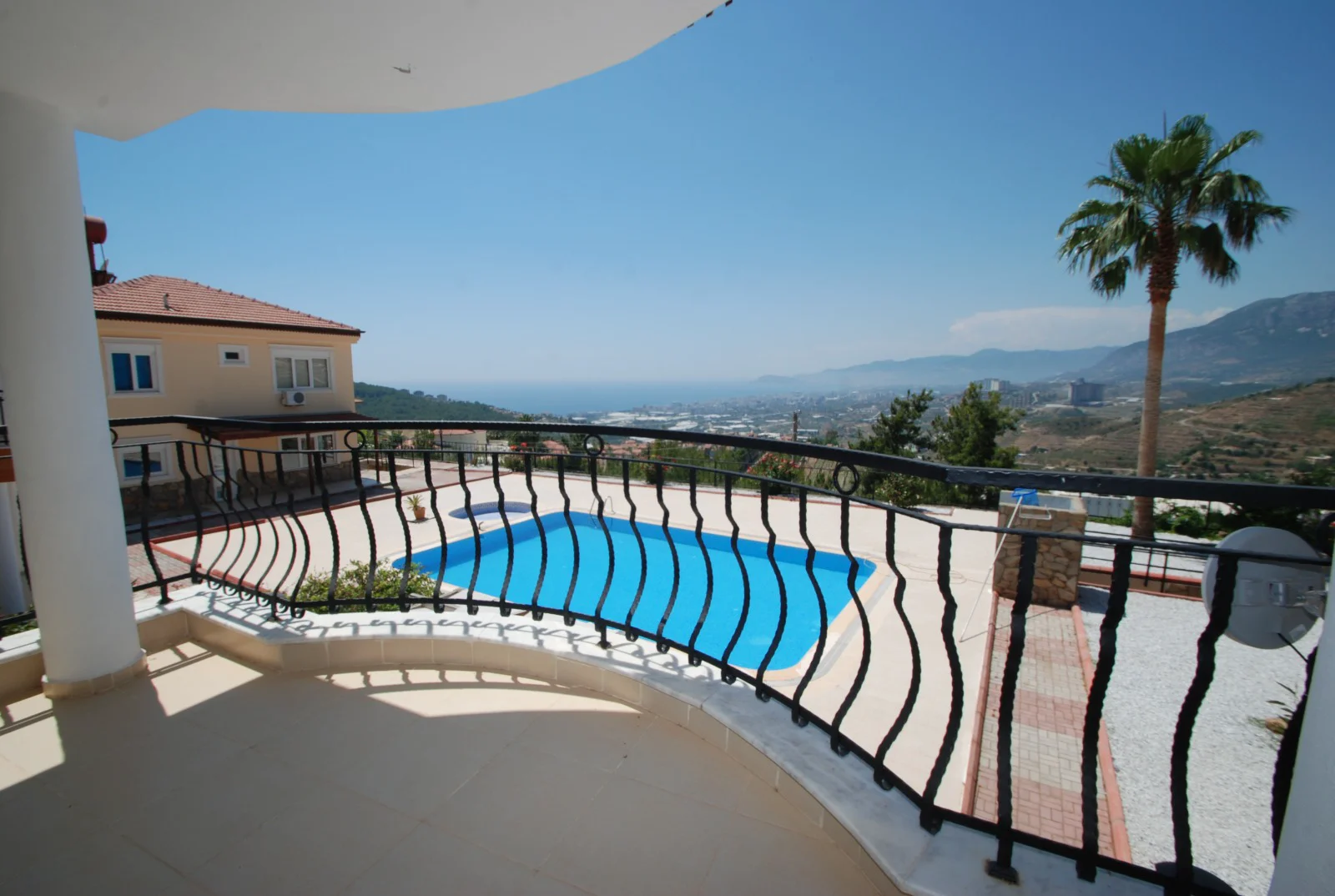 Spacious Balcony with Private Pool and Panoramic Sea Views. A spacious, modern balcony with light tiles and a curved black railing. It overlooks a bright blue swimming pool, offering panoramic views of the sea, city, and mountains.