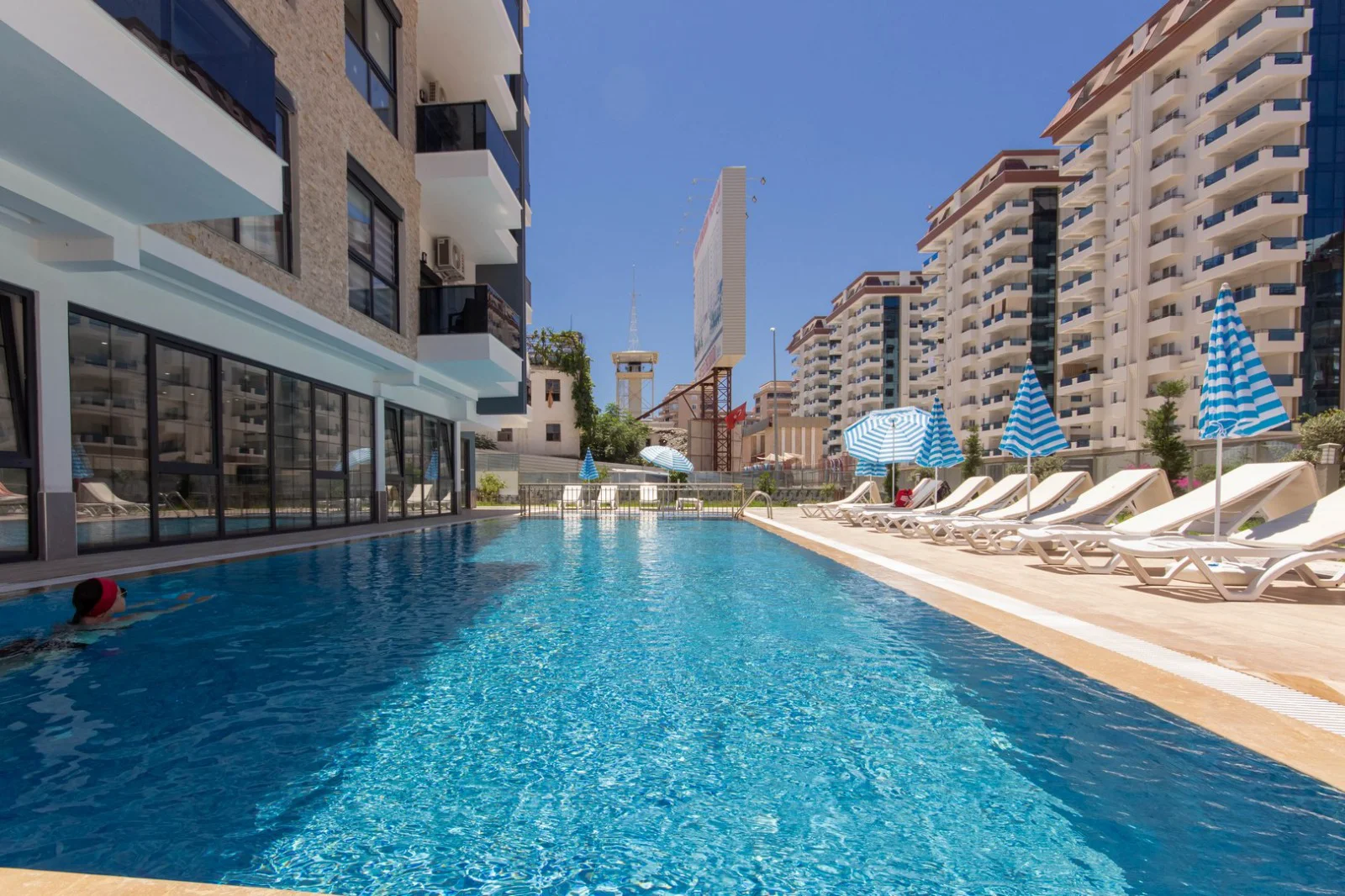 Resort-Style Outdoor Pool with City View. A refreshing outdoor swimming pool area in a modern apartment complex. Features a long rectangular pool, sun loungers with blue striped umbrellas, and multi-story buildings under a clear sunny sky.