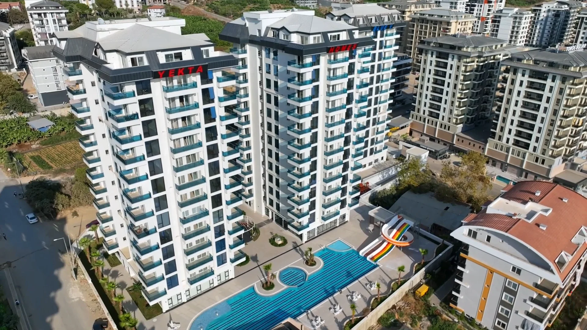 Modern Apartment Complex with Large Pool and Waterslide. Aerial view of a modern residential complex featuring two high-rise apartment buildings, a large outdoor swimming pool with a colorful waterslide, and landscaped grounds.