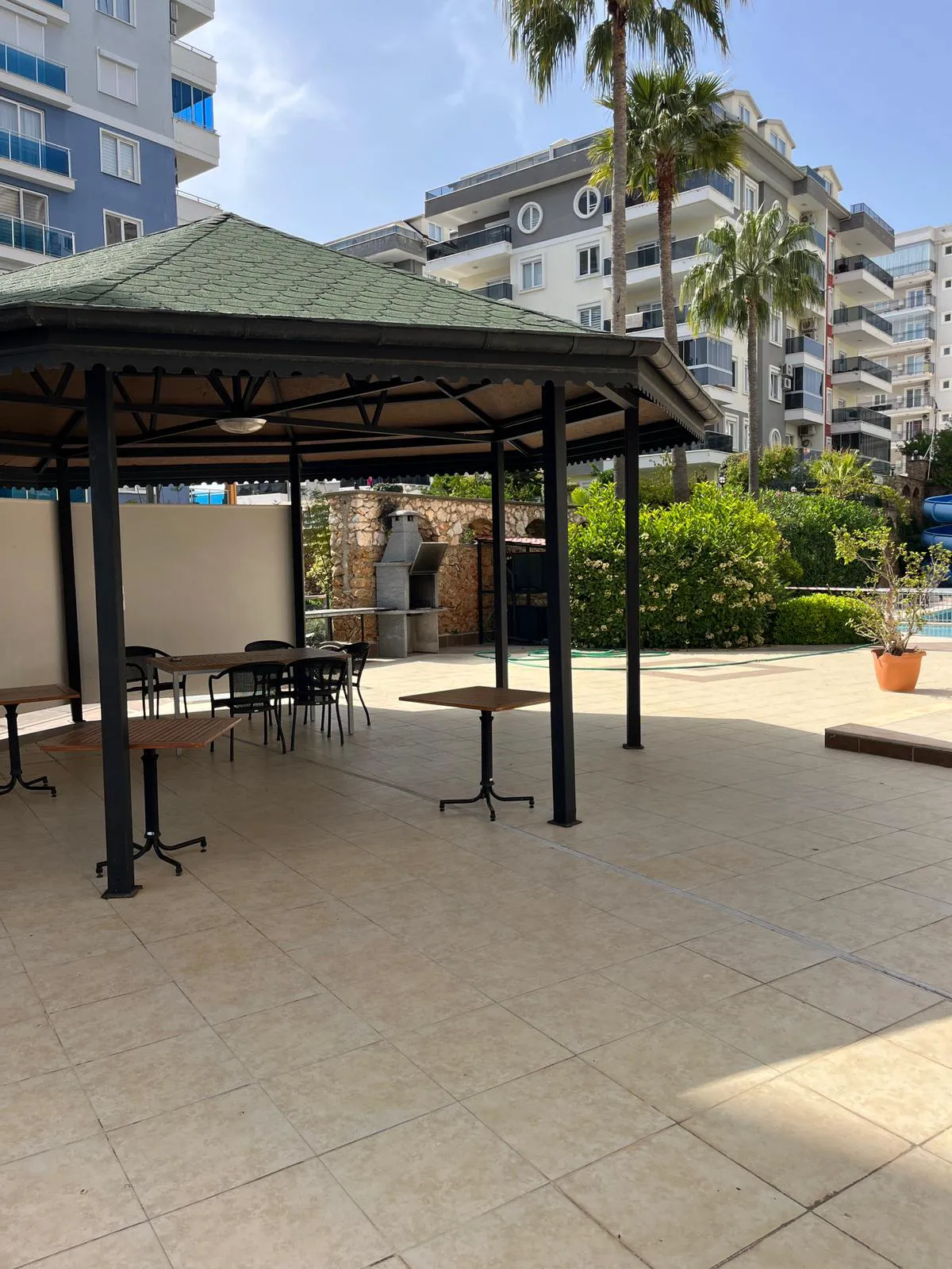 Outdoor Social Area with Gazebo and Barbecue. A well-maintained outdoor communal area featuring a green-roofed gazebo with tables and chairs, a stone barbecue, and lush greenery. Tiled floor leads to a pool area and modern apartment buildings.