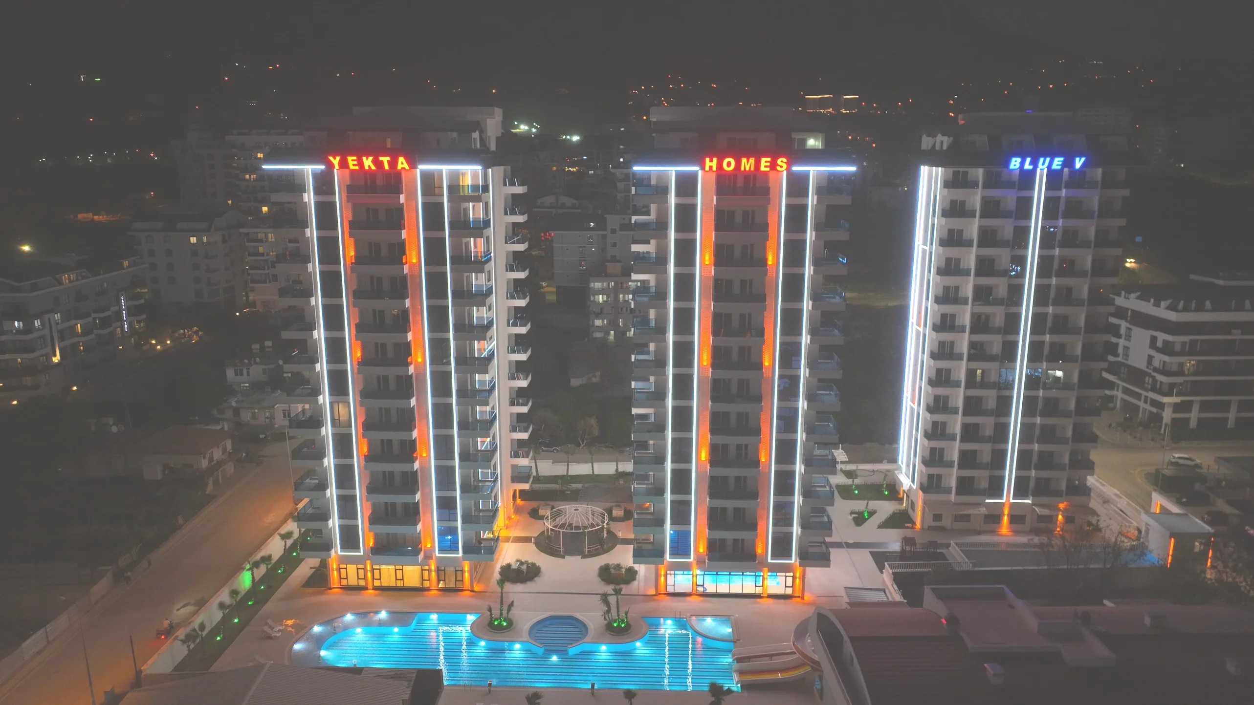 Luxury Apartment Complex with Illuminated Buildings and Pool at Night. Aerial view of a modern luxury apartment complex at night. Three tall buildings are brightly illuminated, featuring an expansive outdoor swimming pool with blue lights and surrounding landscaped green areas, highlighting the property's amenities.