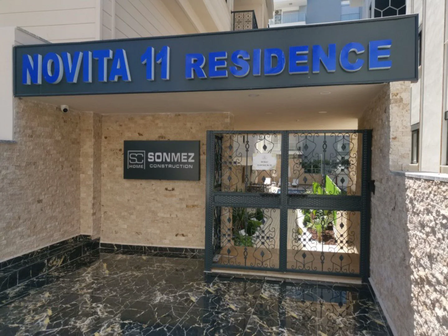 Elegant Entrance to Novita 11 Residence. The modern entrance to Novita 11 Residence building features glossy black marble flooring, light stone walls, and an ornate dark metal security gate. The building name is clearly displayed.