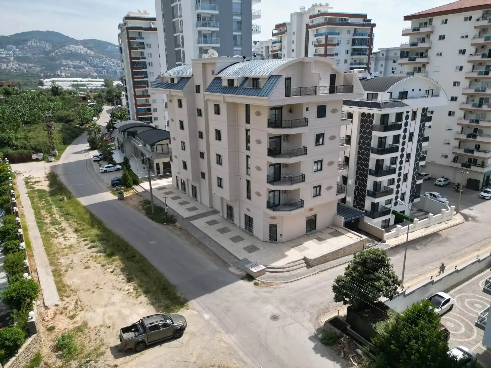 Modern Apartment Building Exterior with City and Hill Views. Aerial view of a modern, multi-story apartment building exterior featuring balconies, situated on a street corner amidst other residential structures, with lush green hills in the background.
