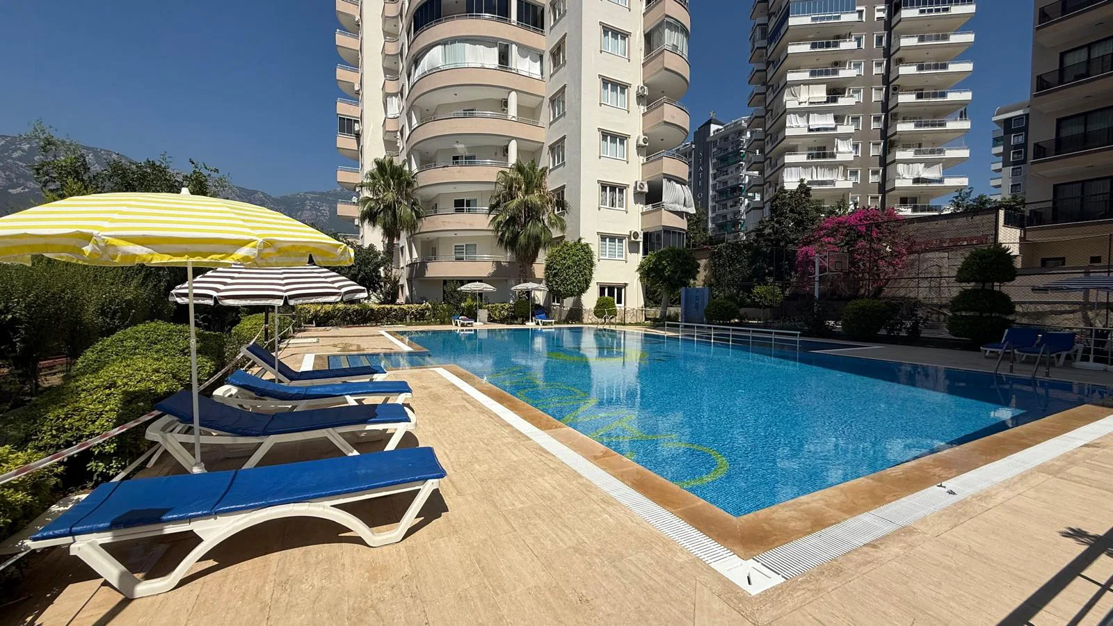 Sparkling Outdoor Swimming Pool in Residential Complex. Vibrant outdoor swimming pool area. Features a large blue pool, sun loungers, yellow umbrellas, and green landscaping. Modern buildings and mountains are visible on a sunny day.