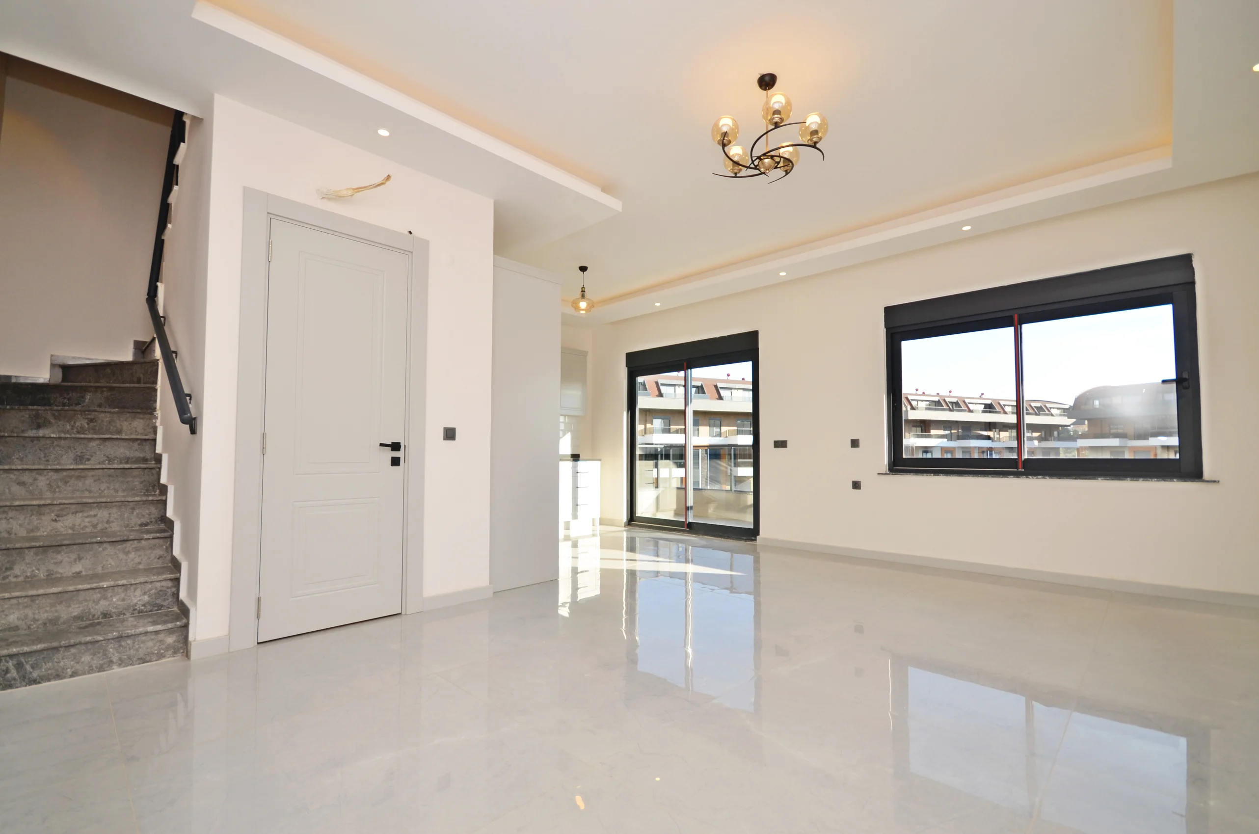 Spacious Modern Living Room with Bright Tiled Floors. A bright, unfurnished modern living room featuring glossy light gray tiled floors, large windows, and a sliding door. The room has recessed lighting, a decorative chandelier, and an adjacent staircase.