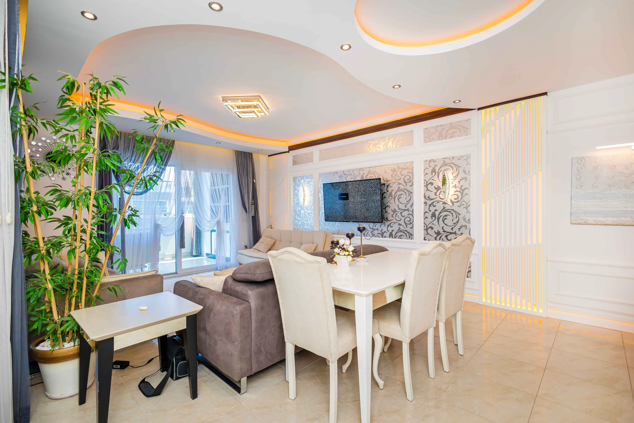 Spacious Modern Living Room with Elegant Dining Area. Modern and elegant living and dining room with light tiled floors, decorative wall panels, and warm cove lighting. Features a dining set, comfortable sofa, wall-mounted TV, and balcony access.