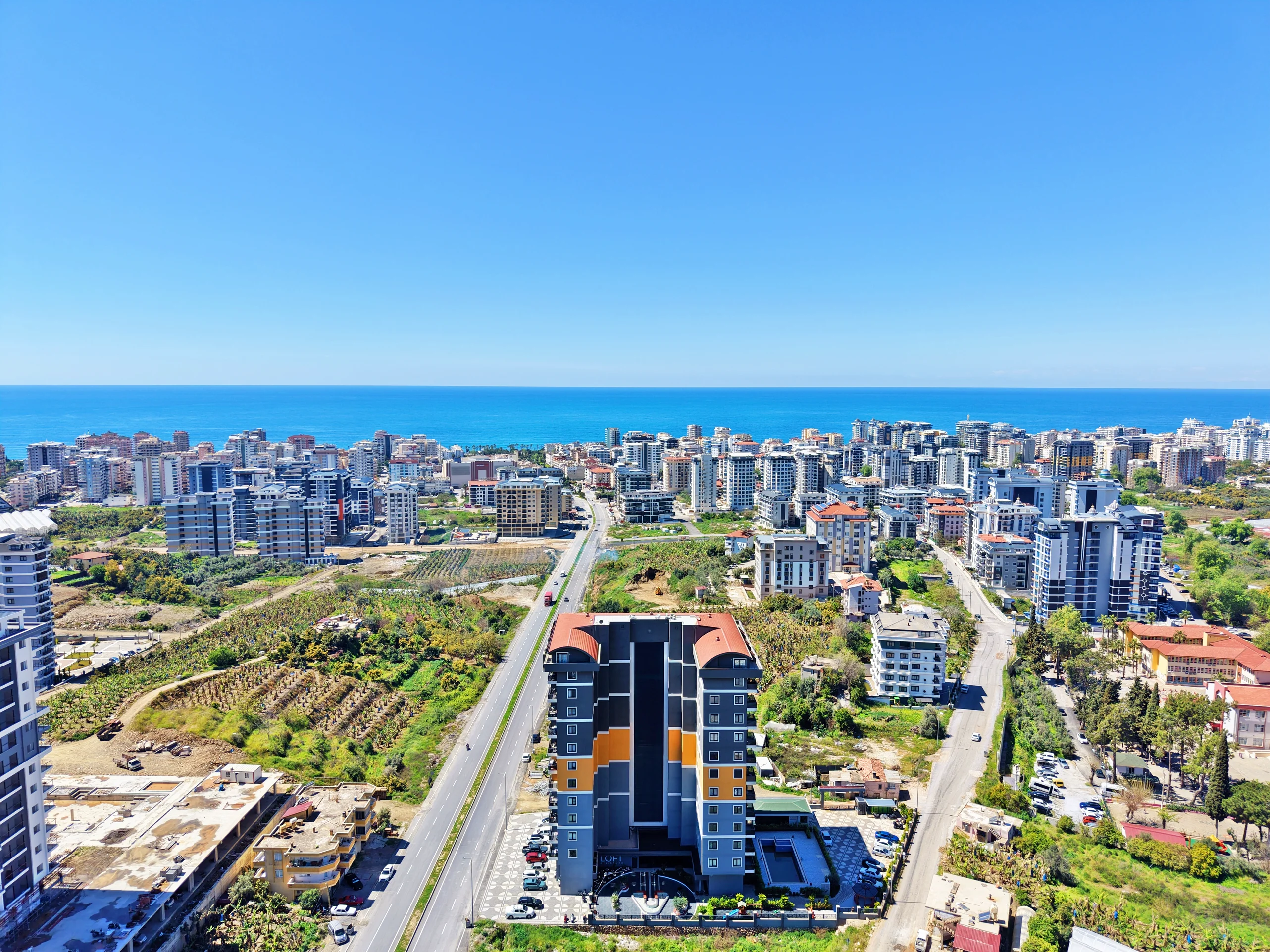 Panoramic Coastal City and Sea View in Turkey. Aerial panoramic view of a vibrant coastal city in Turkey, featuring modern high-rise buildings, green areas, a central road, and the vast blue Mediterranean Sea under a clear sky.