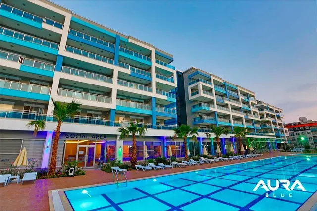 Modern Apartment Complex with Large Outdoor Swimming Pool. Modern apartment complex with a large blue outdoor swimming pool and sun loungers. The building features blue-accented balconies and tropical palm trees, creating a vibrant resort-style ambiance.