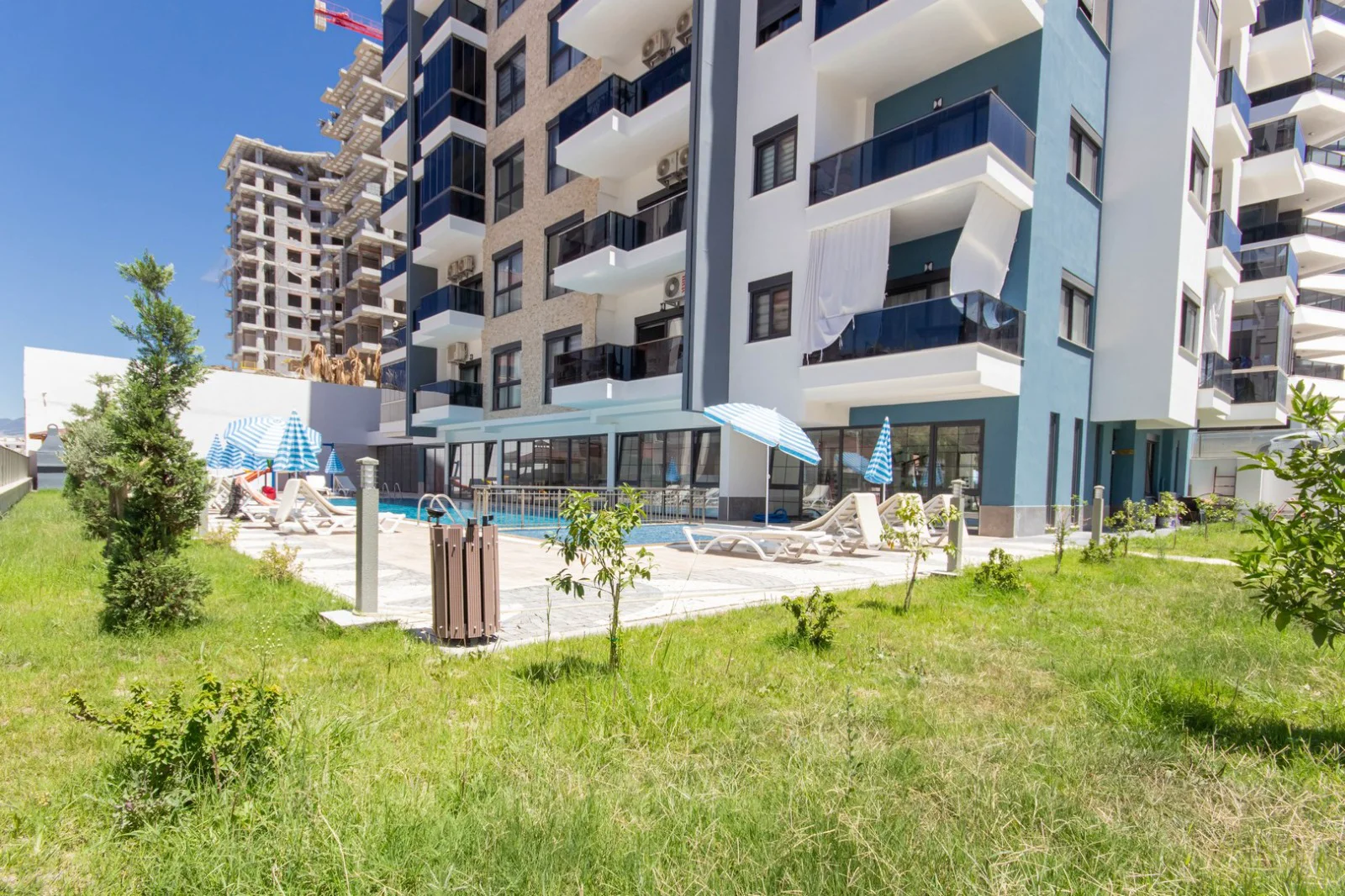 Modern Apartment Complex with Outdoor Swimming Pool and Garden. Modern apartment complex exterior featuring a refreshing outdoor swimming pool with sun loungers and blue umbrellas. A well-maintained green lawn and balconies are also visible.