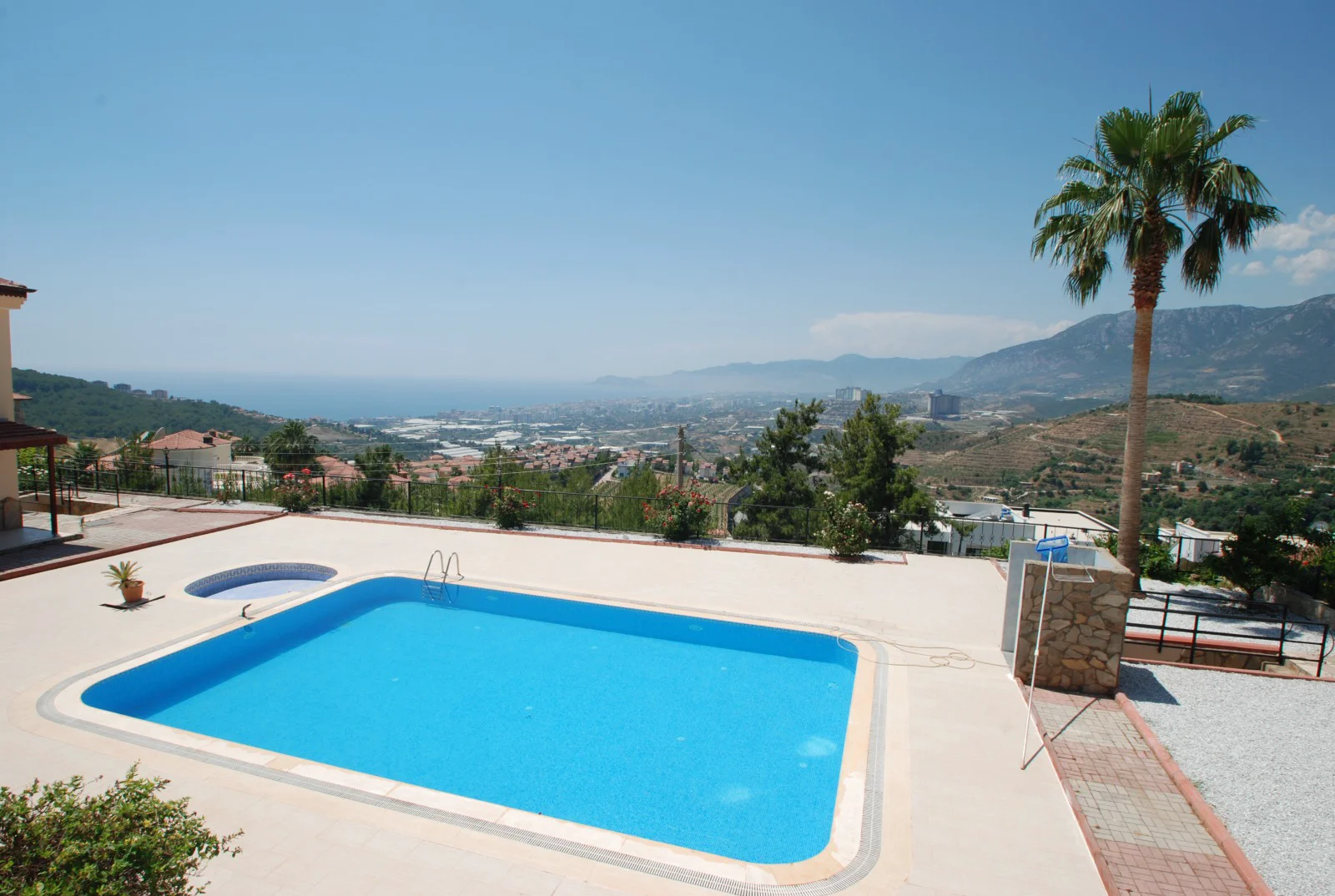 Stunning Panoramic Sea and Mountain Views from Poolside. Outdoor pool area with a modern blue swimming pool and jacuzzi. Offers breathtaking panoramic views of the Mediterranean Sea, coastal city, and distant mountains under a clear blue sky.