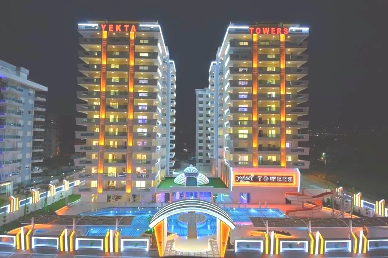 Stunning Night View of Modern Residential Complex with Pool. Exterior view of Yekta Towers, a modern luxury residential complex at night. Two illuminated high-rise buildings overlook a vibrant outdoor pool area with a water slide and colorful accent lighting.