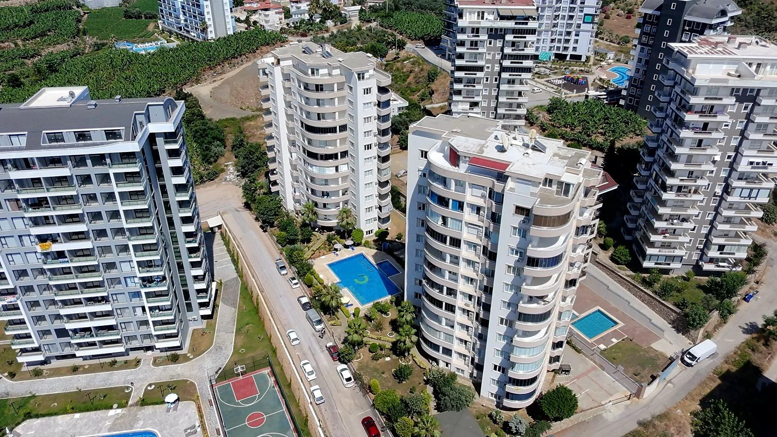 Aerial View: Modern Residential Complex with Pools & Amenities. An aerial perspective of a modern residential complex in Turkey, showcasing several high-rise apartment buildings, vibrant blue swimming pools, a basketball court, and lush green landscaping.