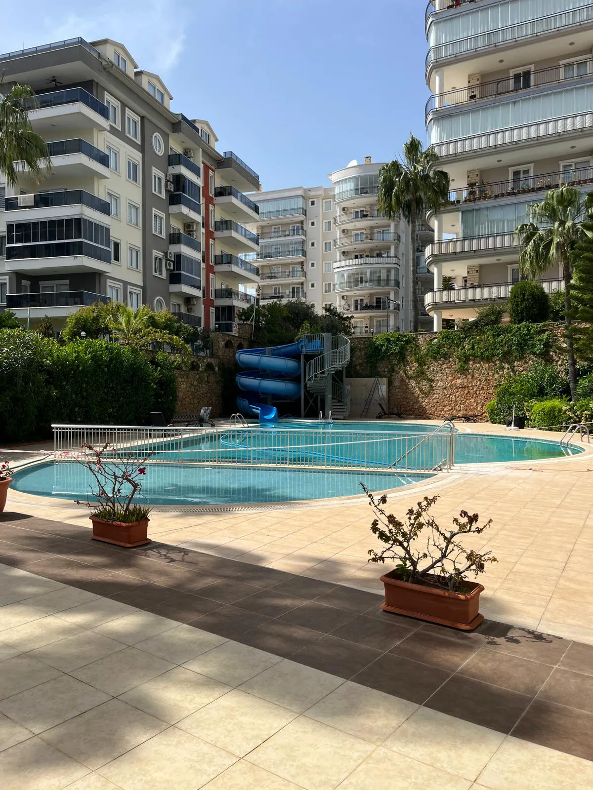 Modern Residential Complex Pool Area with Water Slide. Modern residential complex outdoor pool area with a large swimming pool and a blue water slide. Tiled patio, green landscaping, and apartment buildings create a refreshing leisure space.