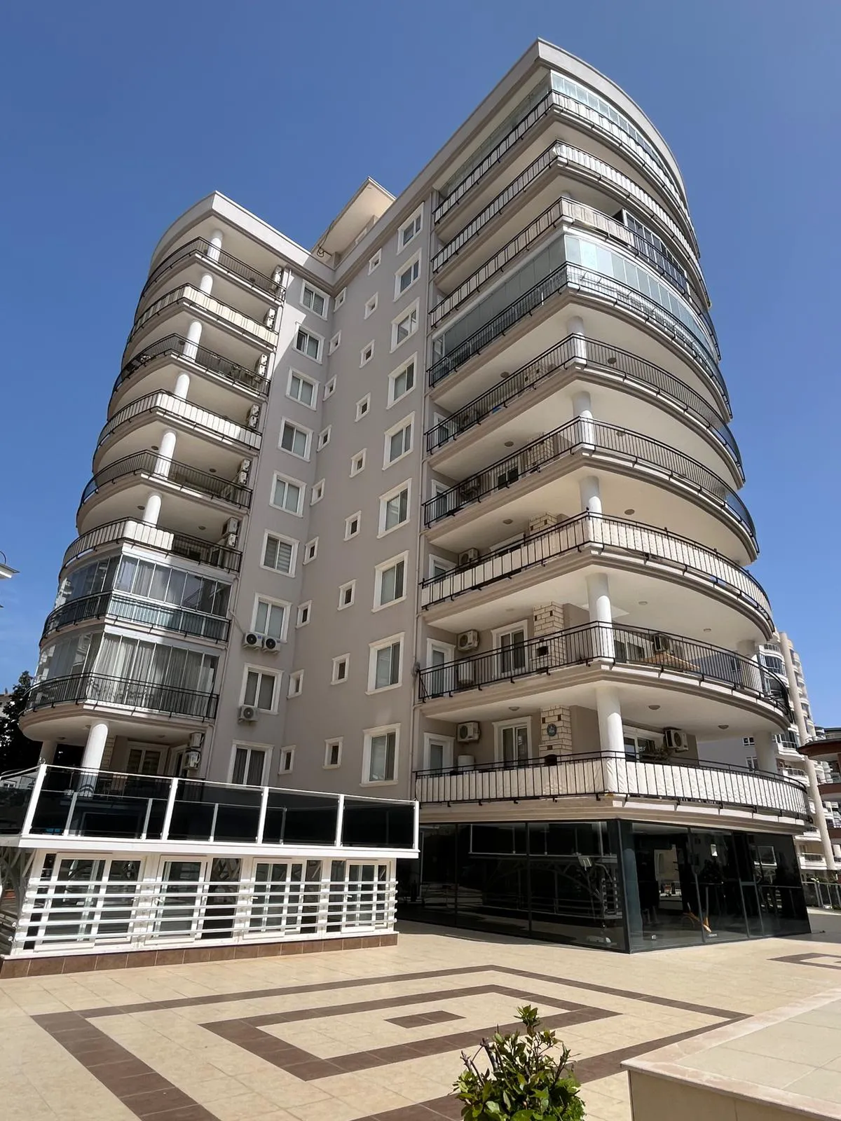 Modern Apartment Building with Curved Balconies Exterior. Exterior view of a modern multi-story apartment building under a clear blue sky. It features a light grey facade with numerous curved balconies, some enclosed with glass, and a tiled plaza in the foreground.