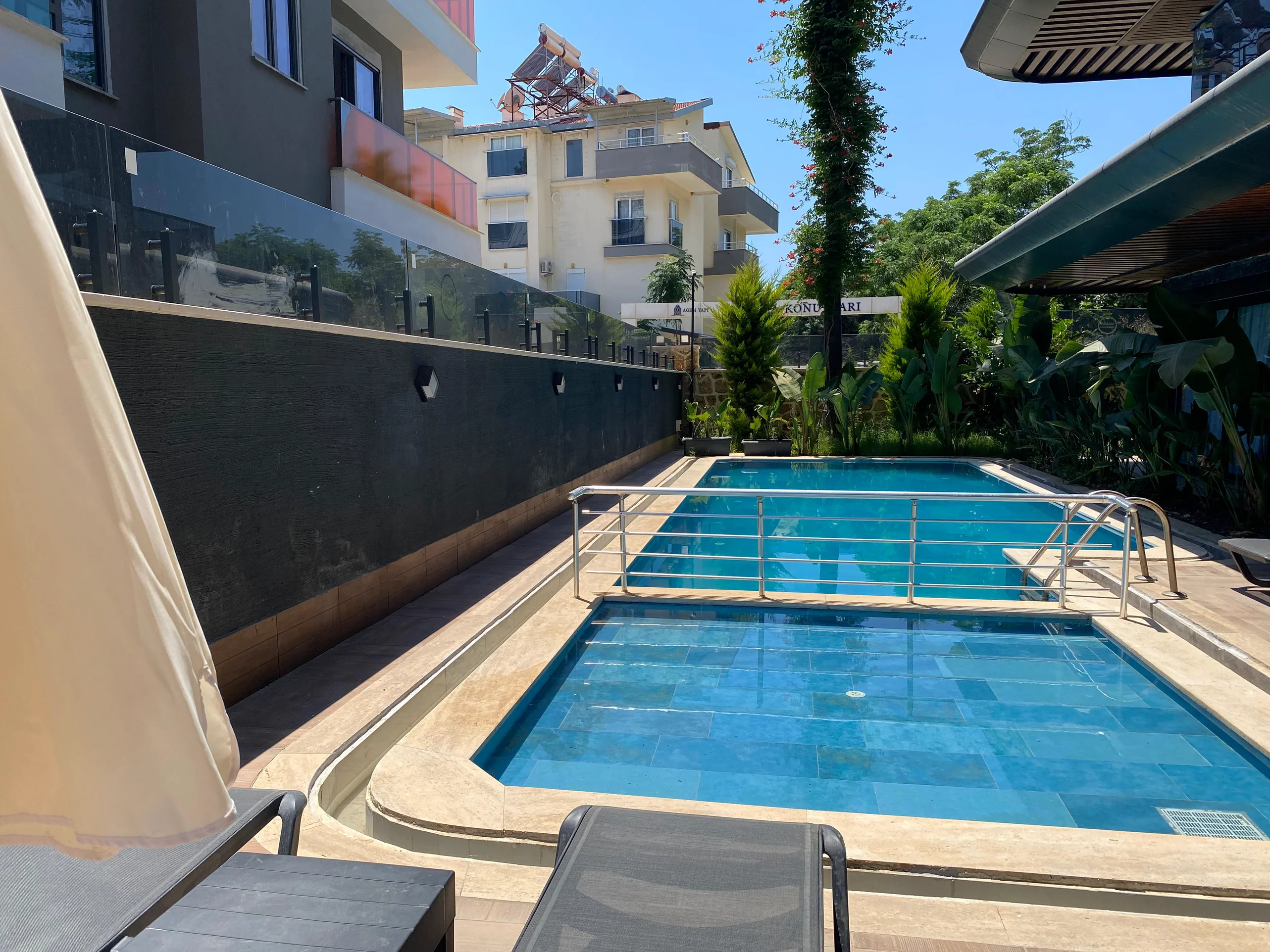 Sunny Outdoor Swimming Pool Area with Greenery. An inviting outdoor pool area featuring two blue-tiled swimming pools with handrails. Sun loungers and a parasol are visible, surrounded by lush greenery. A modern and well-maintained sunny recreational space.