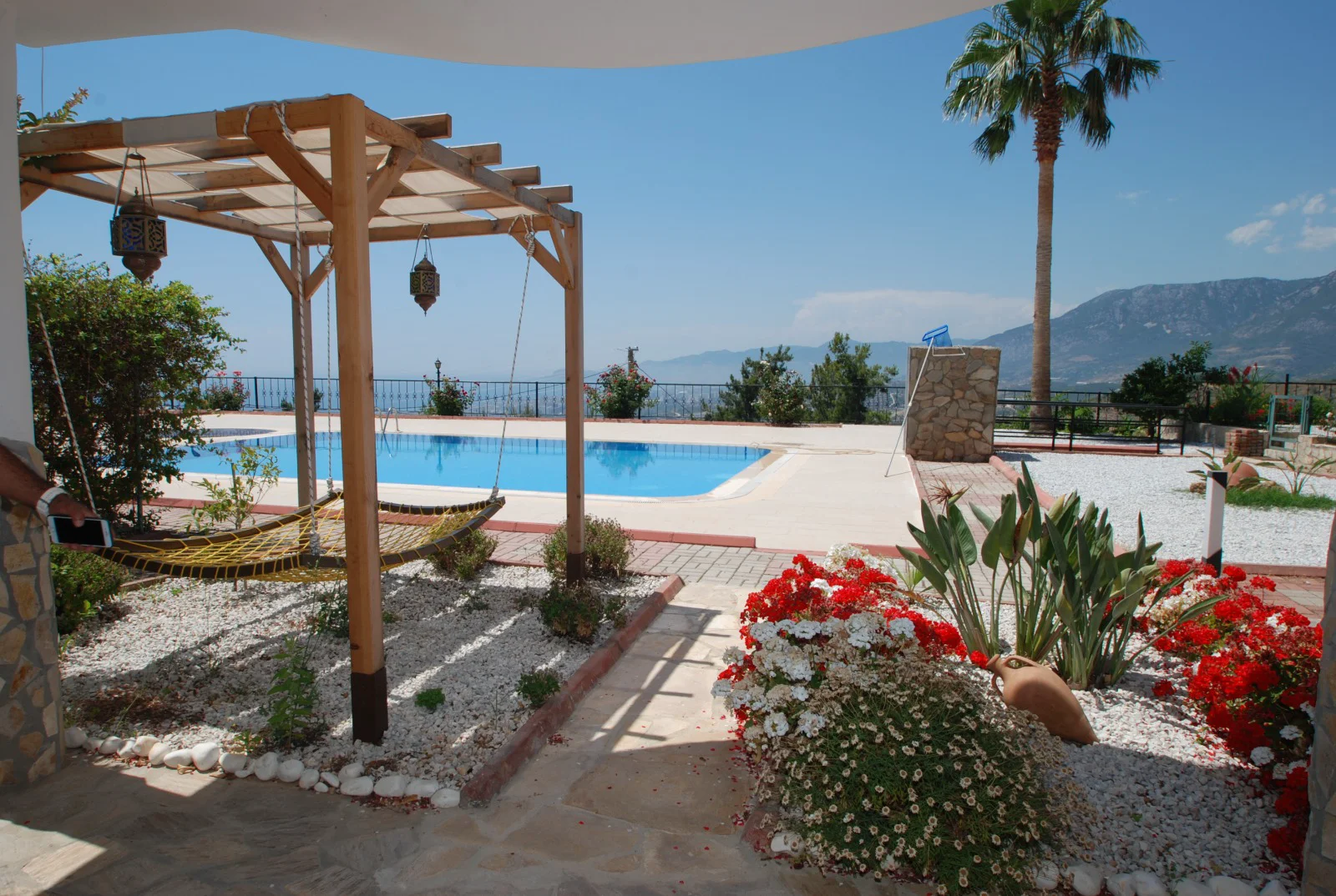 Stunning Outdoor Pool Area with Mountain and Sea Views. A sunny outdoor pool area featuring a sparkling blue swimming pool, a shaded pergola with a hammock, and lush landscaped garden beds with flowers. Offers tranquil mountain views.