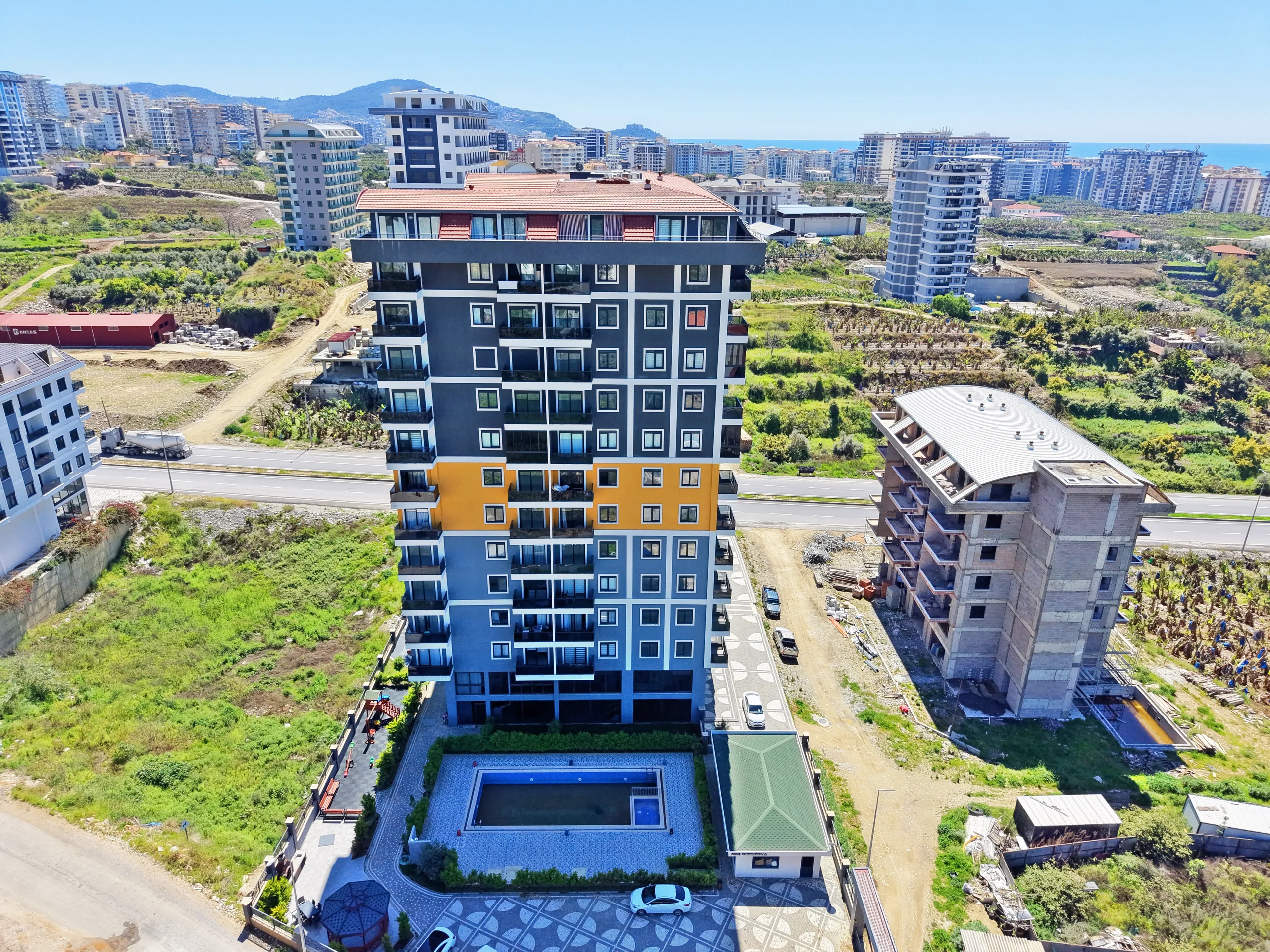 Modern Apartment Building with Outdoor Pool and Playground. Aerial view of a modern multi-story apartment building with an outdoor swimming pool, children's playground, and parking. The contemporary complex offers scenic sea and mountain views.