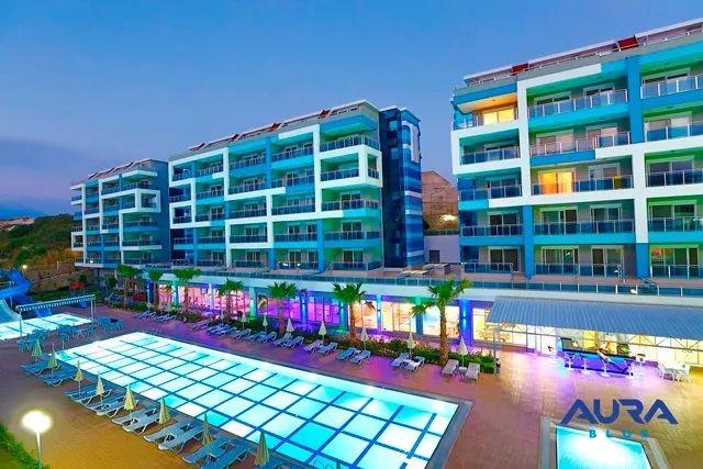 Modern Residential Complex with Vibrant Pools and Resort Amenities. An evening view of a modern resort-style residential complex featuring large, brightly illuminated swimming pools, rows of sun loungers, and contemporary buildings with balconies, surrounded by palm trees and vibrant lighting.