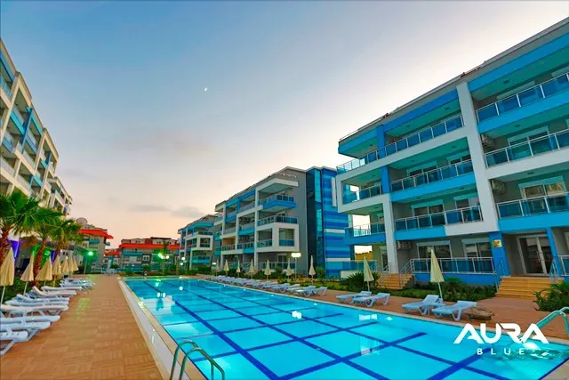 Modern Apartment Complex with Large Outdoor Swimming Pool. Exterior view of a modern apartment complex featuring a large, illuminated outdoor swimming pool, lined with sun loungers and palm trees under a twilight sky.