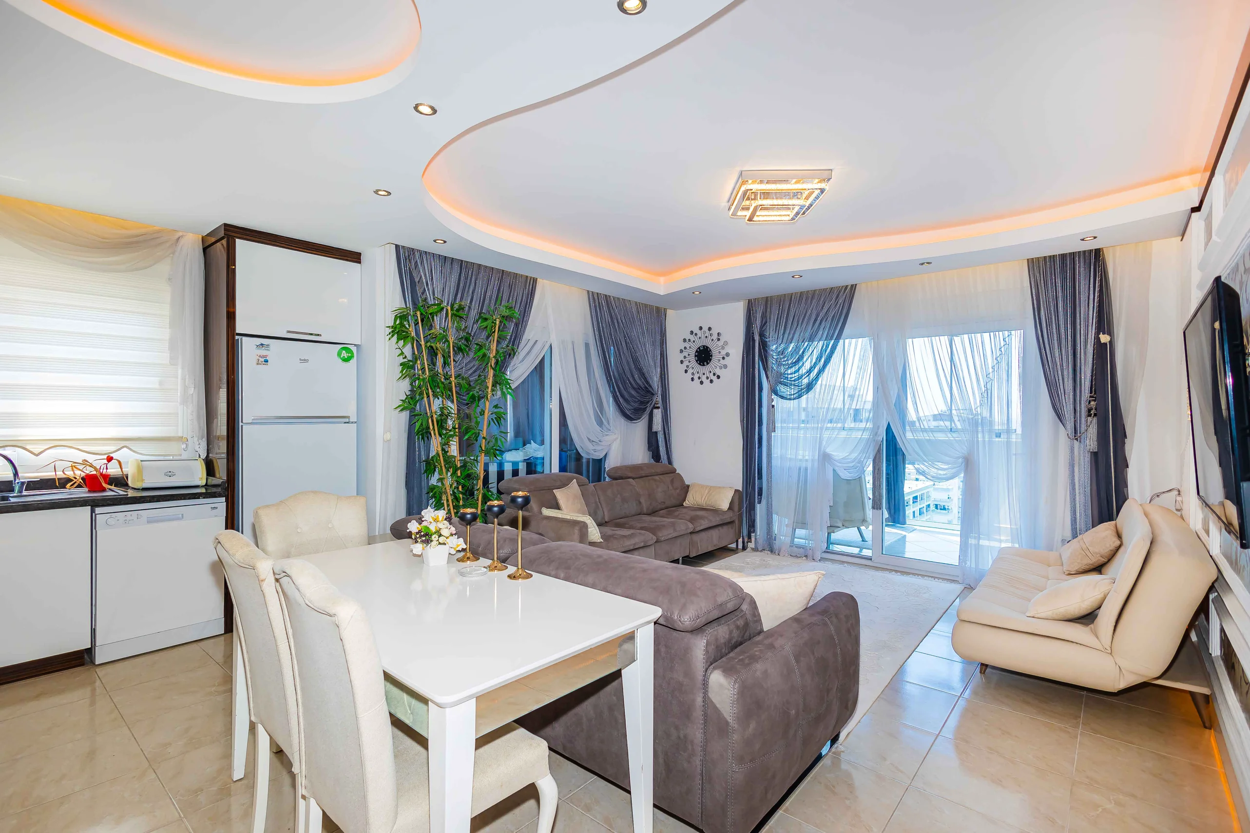 Modern Open-Plan Living Room with Integrated Kitchen and Balcony. Modern open-plan living and kitchen area with tiled floor, contemporary sofas, and dining set. Features integrated appliances, stylish lighting, and glass doors to a balcony.