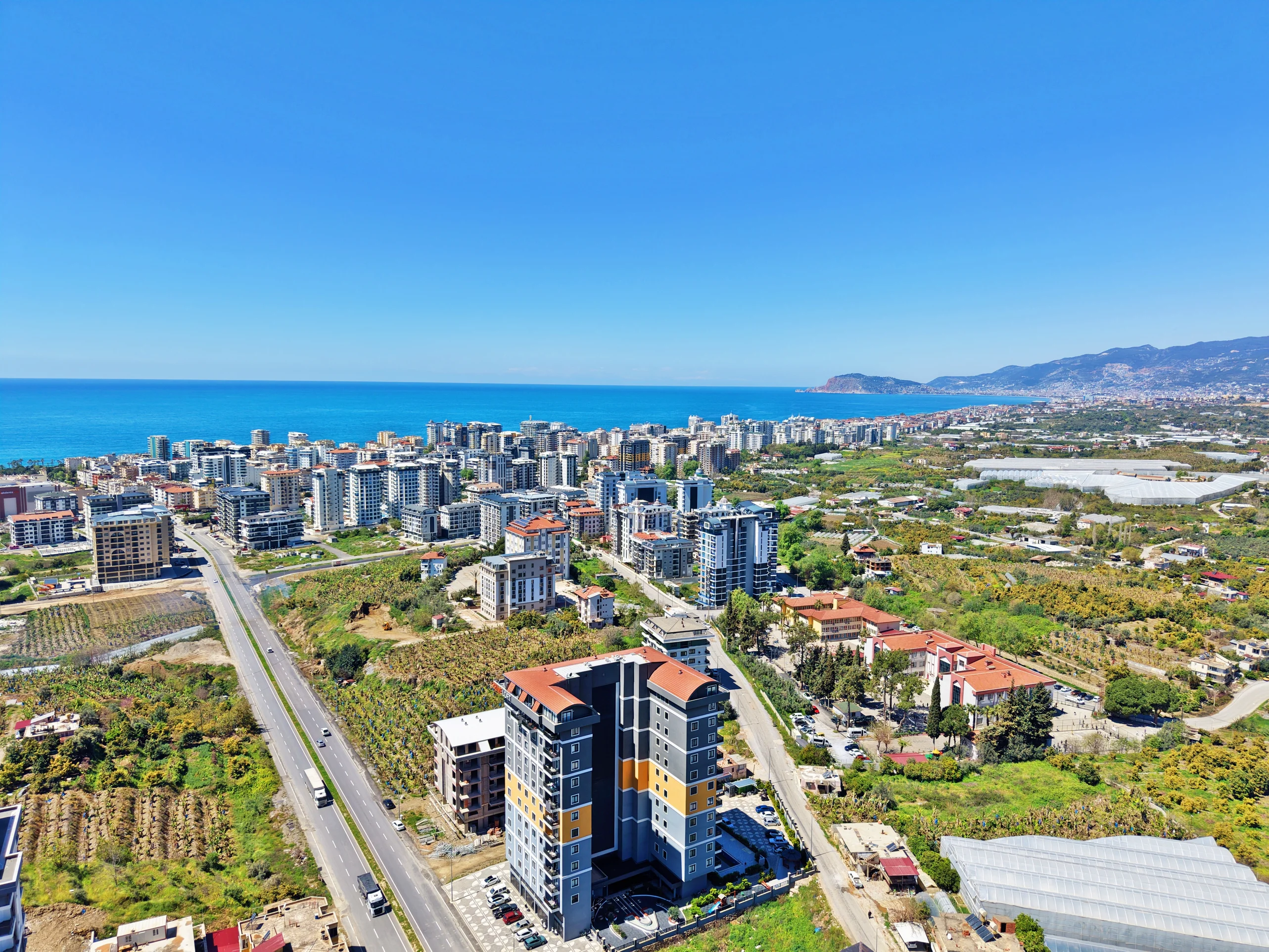 Panoramic Coastal City View with Modern Buildings and Sea. Exterior aerial view of a vibrant coastal city in Turkey, showcasing numerous modern apartment buildings, an azure sea, green agricultural land, and distant mountains under a clear blue sky.