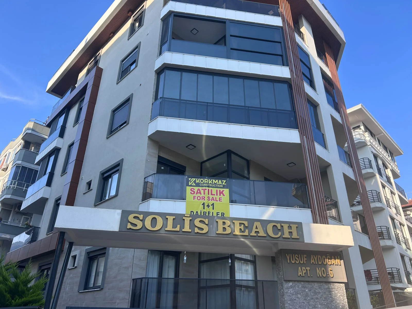 Modern Solis Beach Apartment Building Exterior with Balconies. Exterior view of the modern 'Solis Beach' apartment building featuring multiple floors, contemporary architecture, and glass-railed balconies. A 'For Sale' sign indicates 1+1 apartments are available.