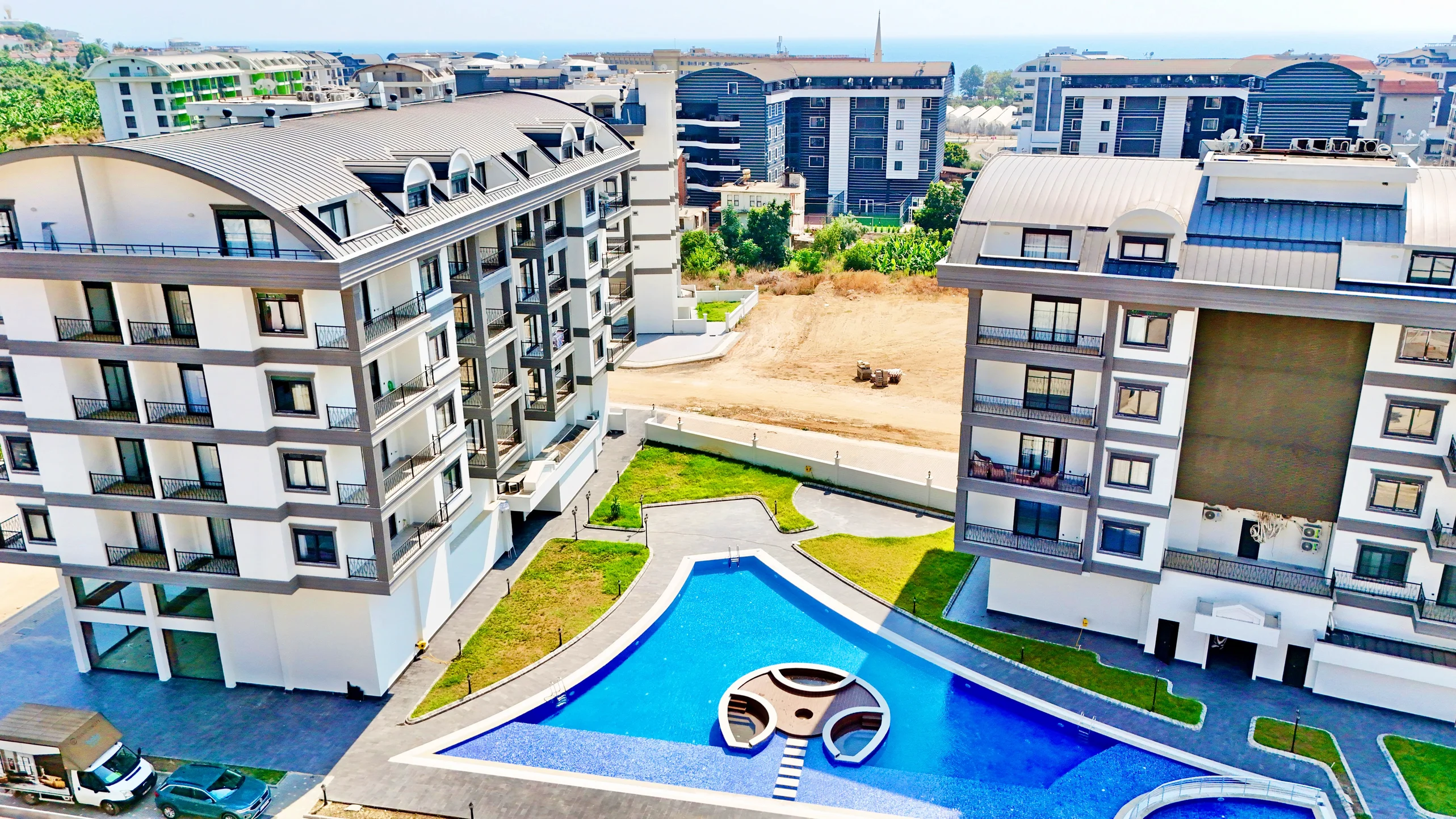 Modern Apartment Buildings with Large Outdoor Pool. Aerial view of a modern residential complex featuring two multi-story apartment buildings, a large blue swimming pool with a unique island, and green landscaped areas, all under a bright sky.