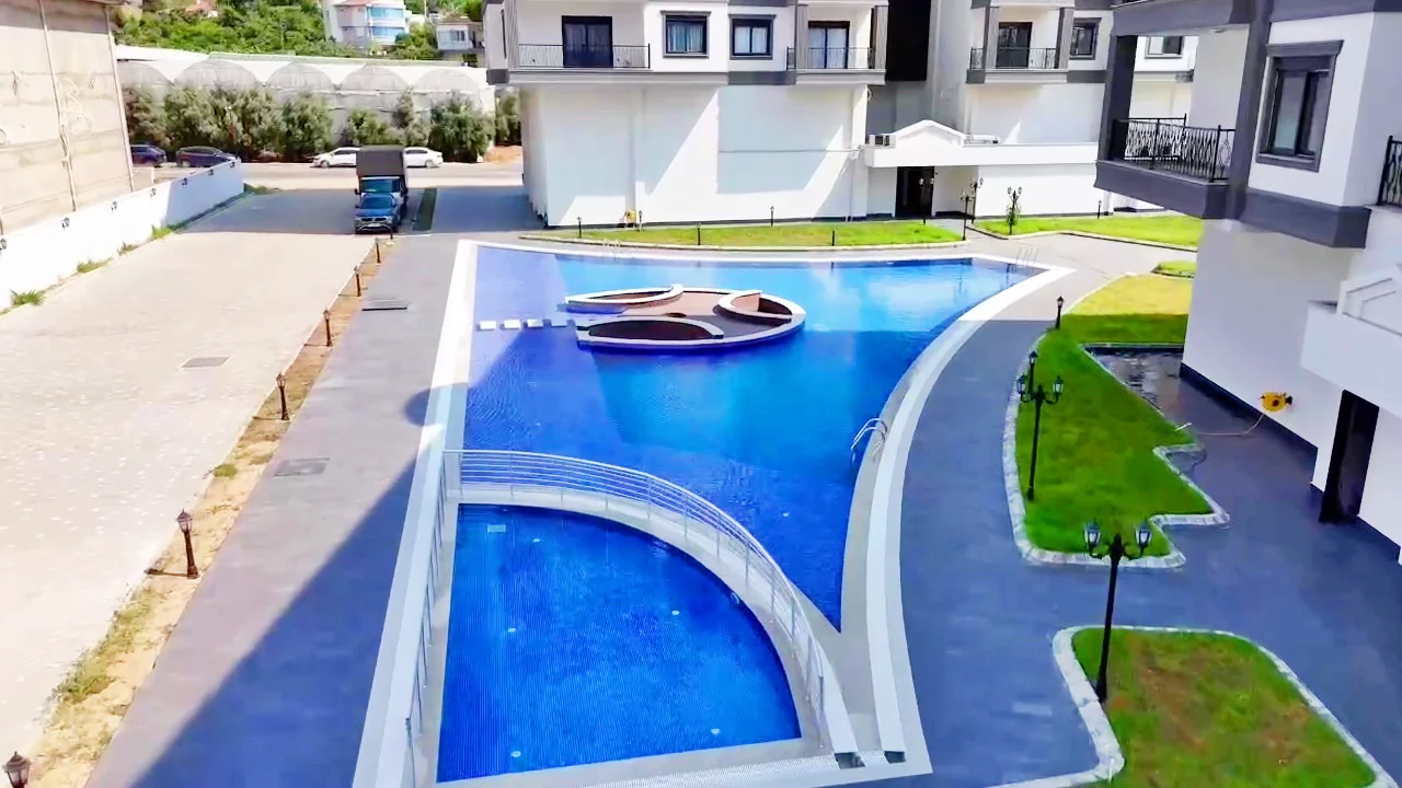 Stunning Outdoor Swimming Pool Area with Modern Design. Aerial view of a modern outdoor swimming pool with a unique floating island and curved bridge. Surrounded by paved walkways, green lawns, and contemporary apartment buildings.