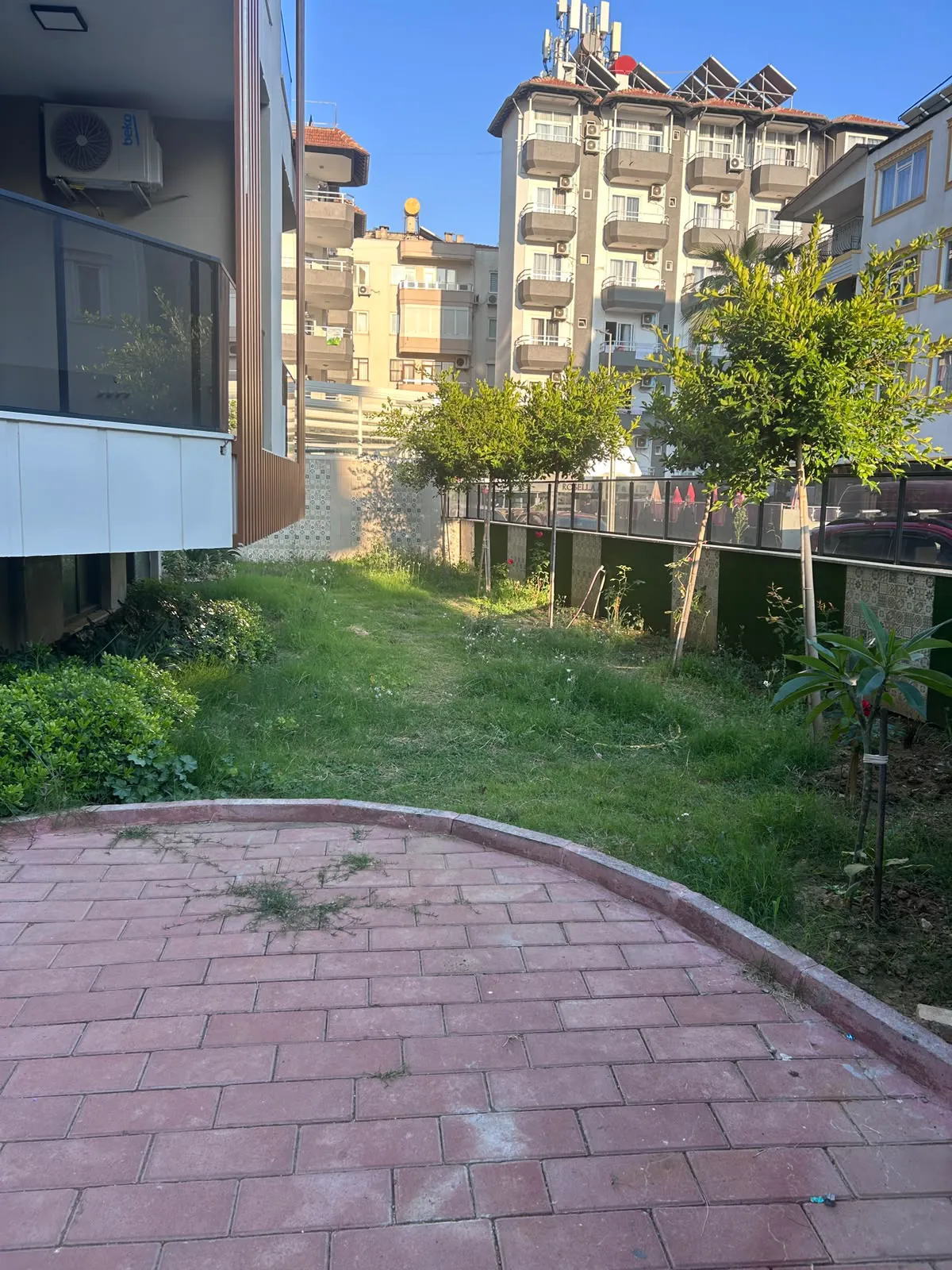 Residential Garden with Paved Patio and Urban Backdrop. An exterior view of a residential garden featuring a red paved patio area and a grassy lawn with young trees, set against apartment buildings under a bright sunny sky.